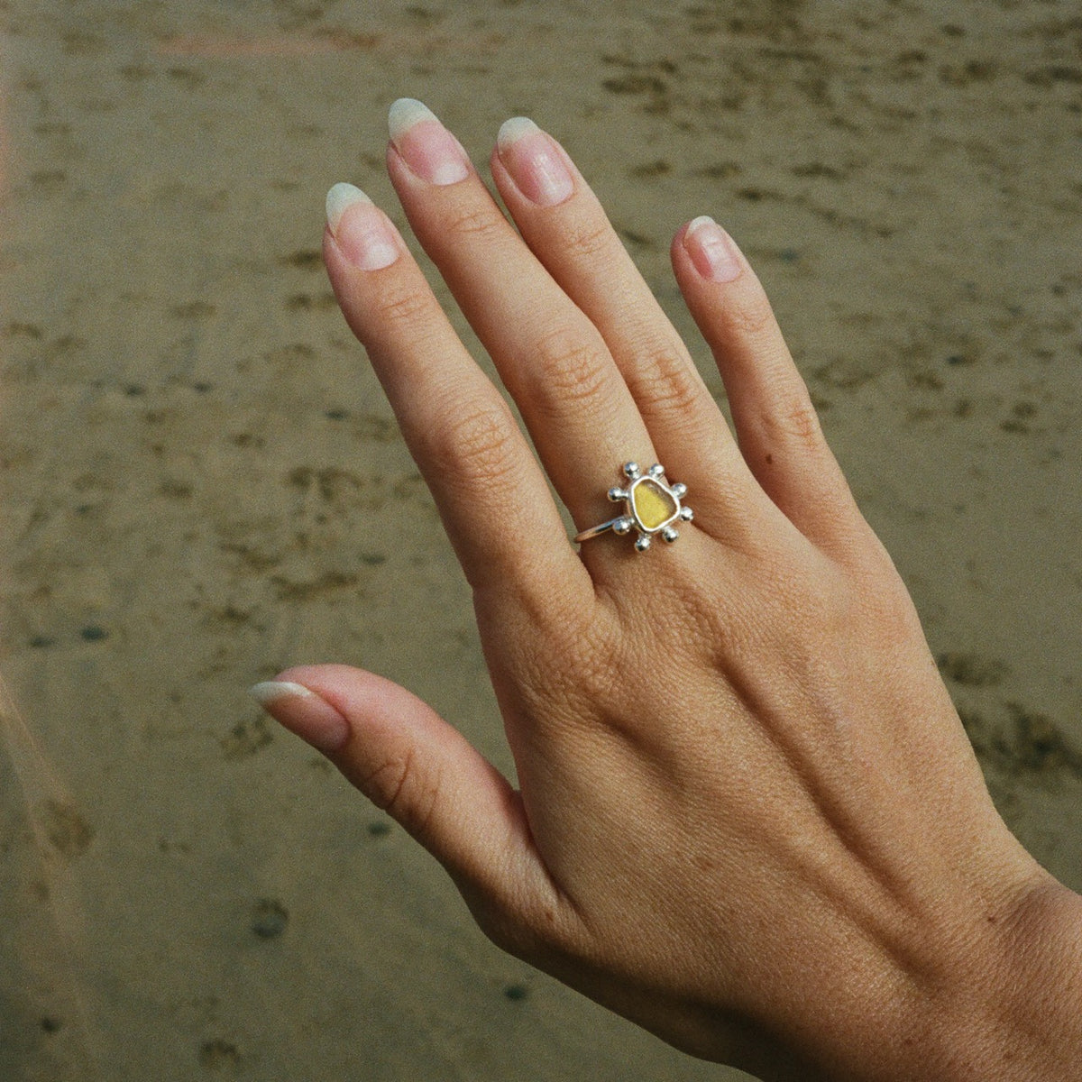 A silver ring with a yellow teardrop-shaped stone and small clear stones around it, worn on a person's ring finger against a sandy background.