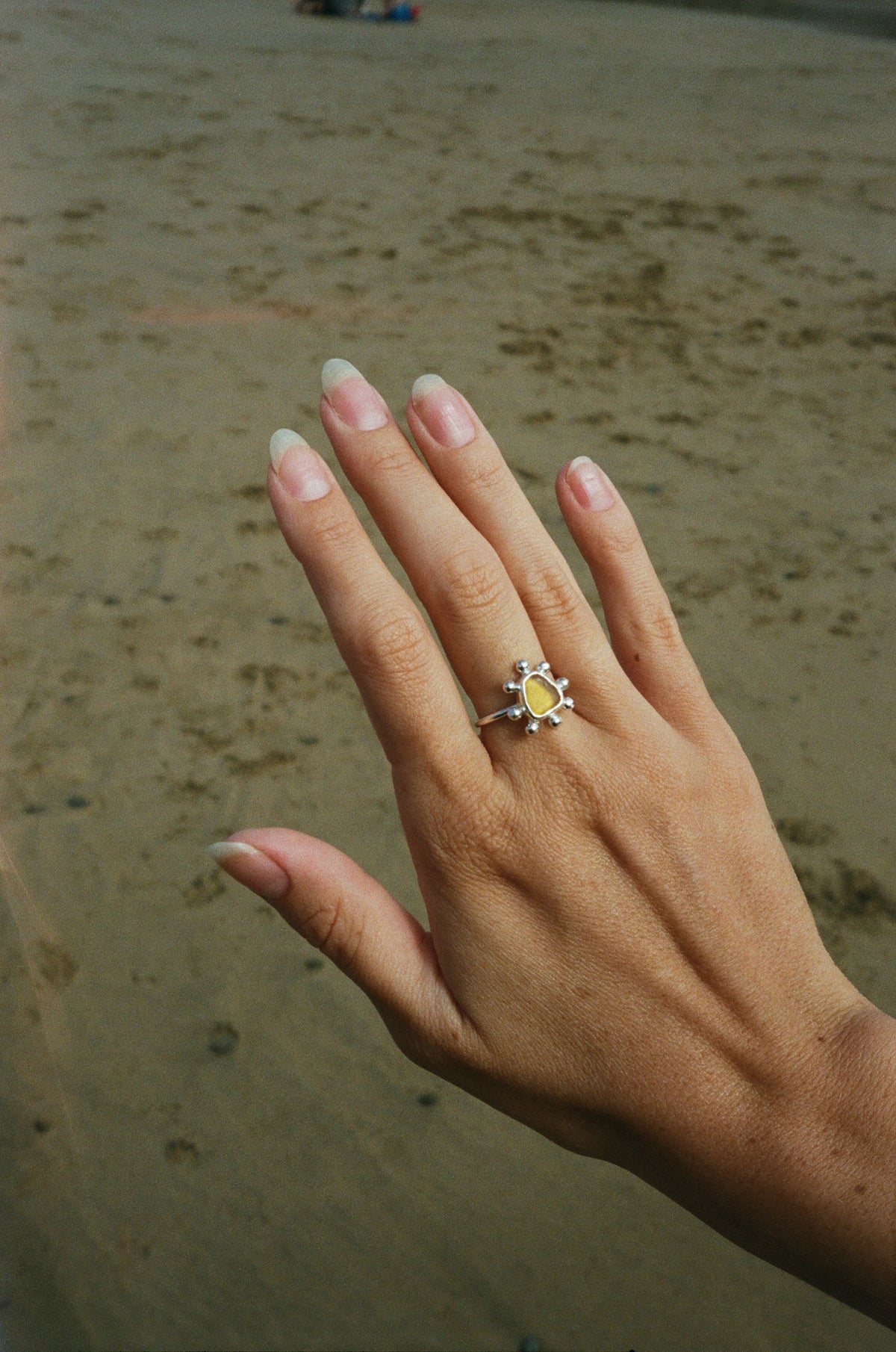 A silver ring with a yellow sunburst design and small blue stones, worn on a hand with a sandy beach background.