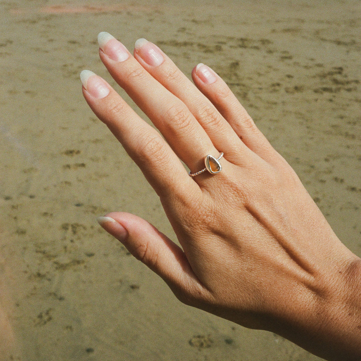 A close-up of a hand with long manicured nails wearing a silver ring with a teardrop-shaped clear stone, set against a blurred sandy beach background.