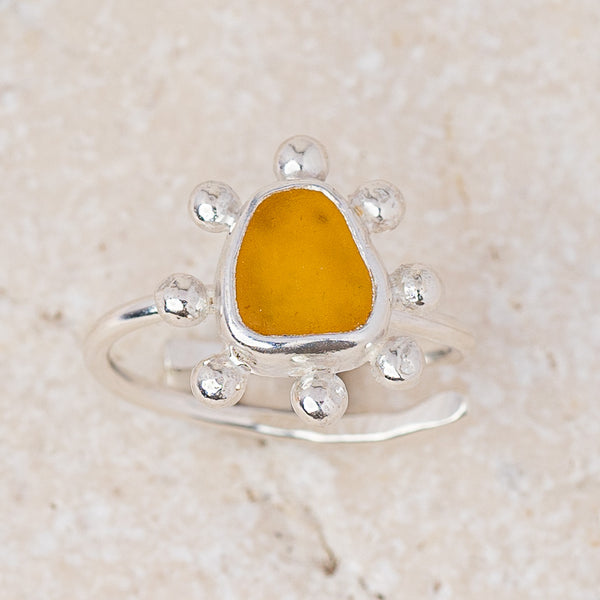 A sterling silver ring featuring a central irregular-shaped yellow amber stone, surrounded by six small silver beads on the band, set against a light beige textured stone background.