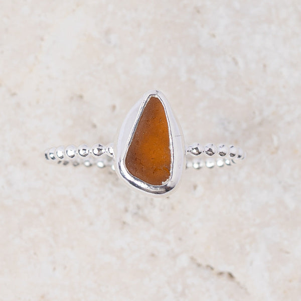 A silver ring featuring a teardrop-shaped amber glass stone, set against a light beige textured stone background.