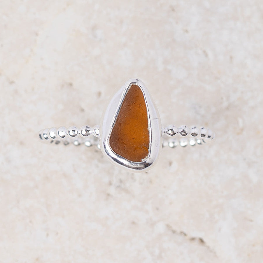 A silver ring featuring a teardrop-shaped amber glass stone, set against a light beige textured stone background.