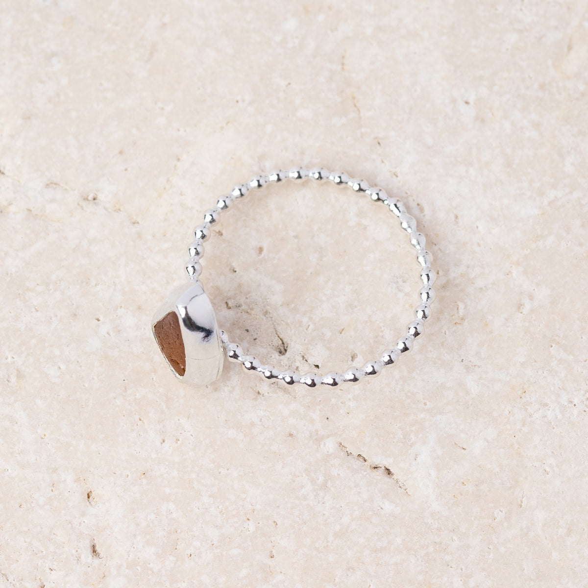 A silver beaded ring with a polished triangular bezel holding a brown gemstone, displayed on a light beige stone surface.