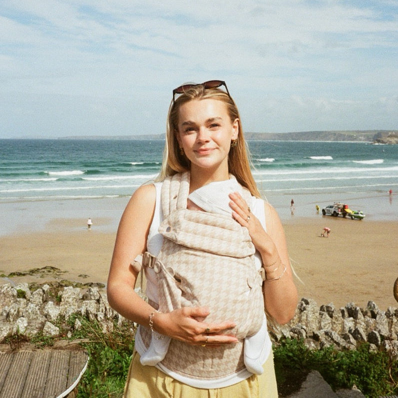 A woman standing on a beach path wearing a beige and white checkered baby carrier wrap with a baby inside, with the ocean and sandy beach in the background.