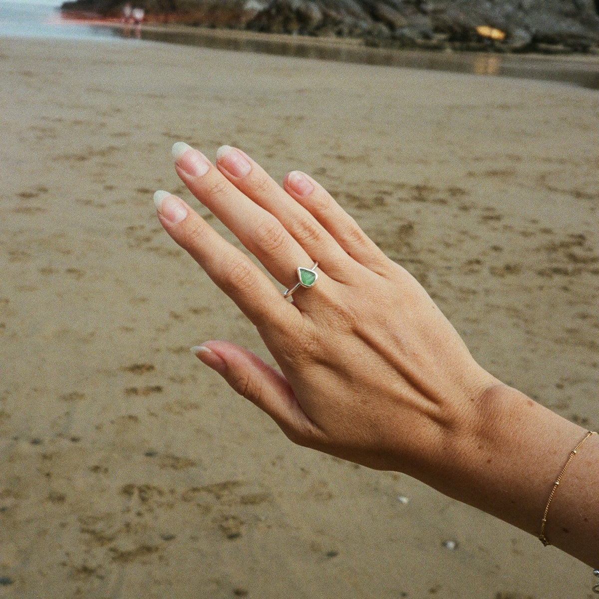 A silver ring with a green teardrop-shaped gemstone worn on a person's hand, set against a blurred sandy beach background.