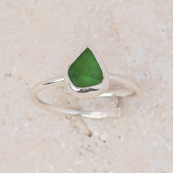 A sterling silver open band ring featuring a triangular bezel-set green sea glass stone, displayed on a light beige textured stone surface.