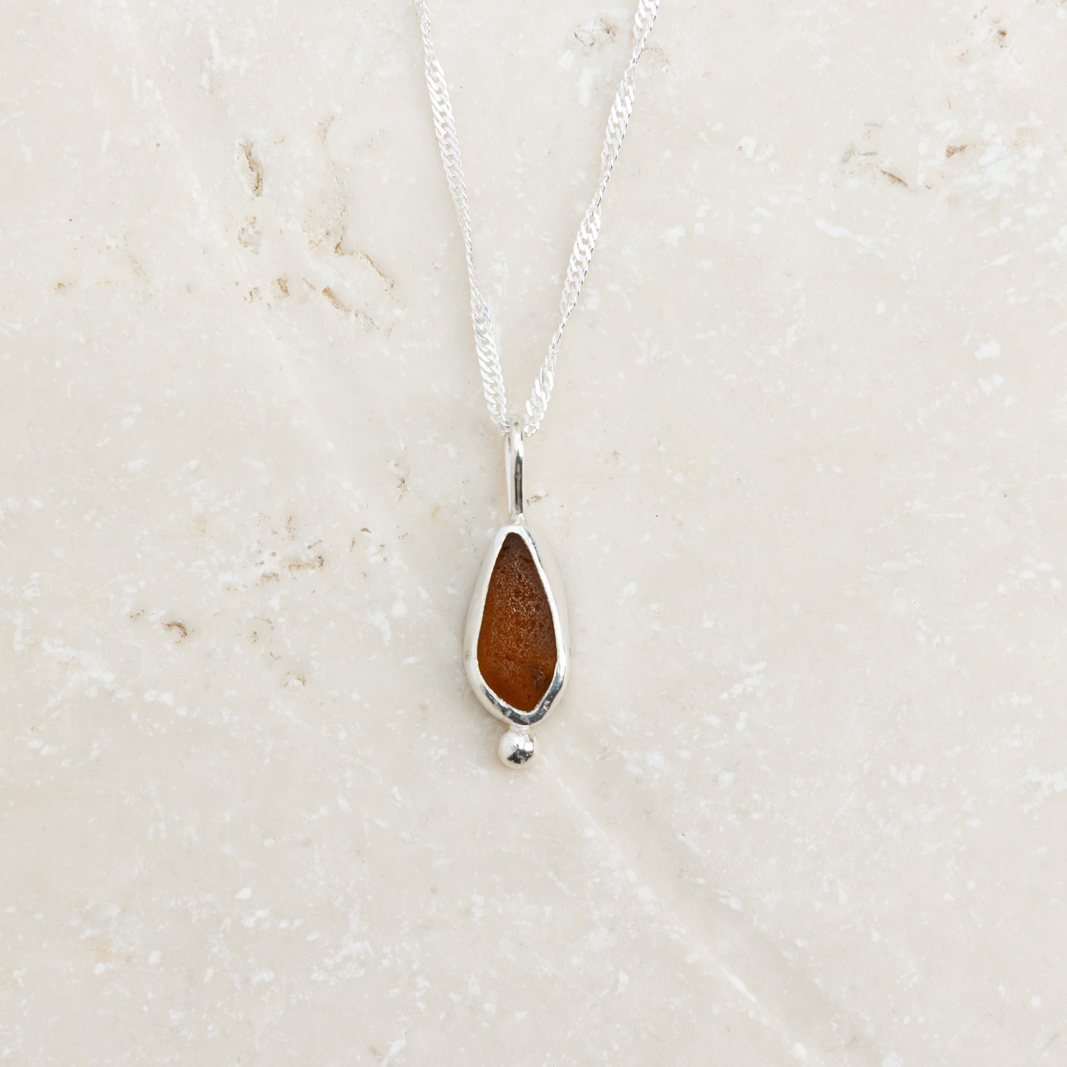 Warm amber sea glass necklace in sterling silver on pale sandy surface, Broken Wave Jewellery Cornwall
