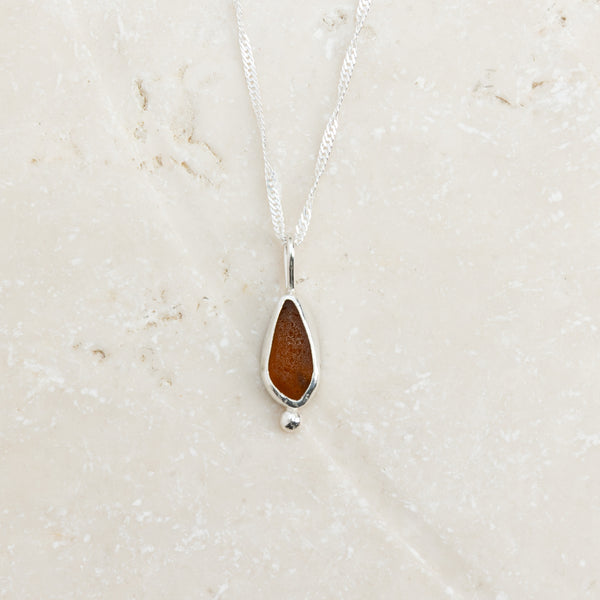 Warm amber sea glass necklace in sterling silver on pale sandy surface, Broken Wave Jewellery Cornwall
