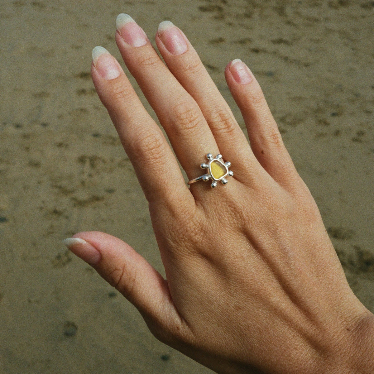 A silver ring with a yellow sunburst design and small blue stones, worn on a hand with a sandy beach background.